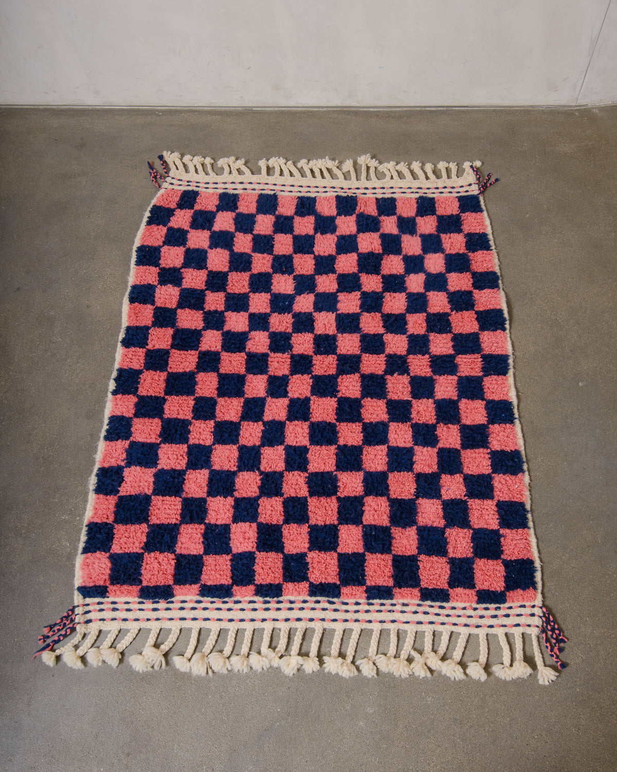 Small Berberlin Beniourain rug made of wool in pink and dark blue checkers, laying on a concrete floor.